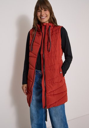 Femme souriante portant une veste matelassée rouge sans manches sur un haut noir à manches longues et un jean bleu, debout contre un mur uni.