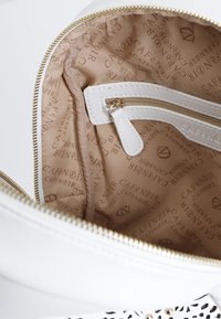 Valkoinen nahkalaukku, jossa on kultainen vetoketju. Laukun sisäpuolella on beige vuori, joka on vuorattu toistuvalla logokuvioinnilla, sekä pieni vetoketjullinen tasku.