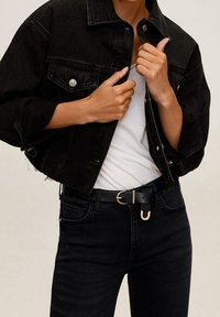 Henkilö, joka on pukeutunut lyhyeen mustaan farkkutakkiin, valkoiseen t-paitaan, mustiin farkkuihin ja mustaan vyöhön, jossa on kultainen solki, pitää takin kauluksesta kiinni.