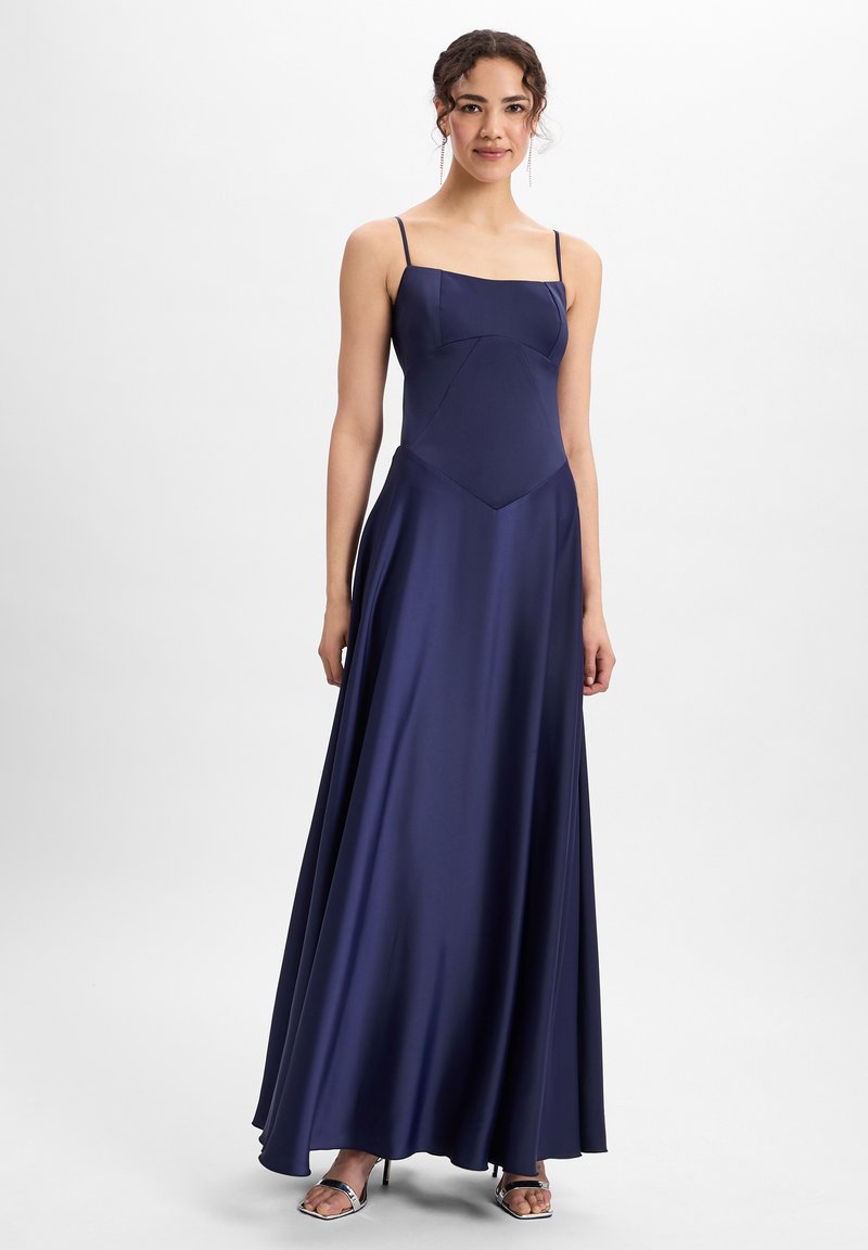 Navy satijnen maxi-jurk met een vierkante halslijn, slanke bandjes en een vloeiende rok. De stof heeft een gladde, glanzende textuur. Zilveren hoge hakken sandalen.