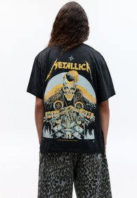 Černé bavlněné tričko s velkým grafickým potiskem lebky s kožešinovými akcenty a kovovými prvky. Tučný text "Metallica" nahoře.