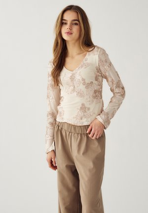 Kobieta z długimi włosami, ubrana w przezroczystą, haftowaną bluzkę w kwiaty i luźne beżowe spodnie, stojąca na tle jasnego, jednolitego tła.