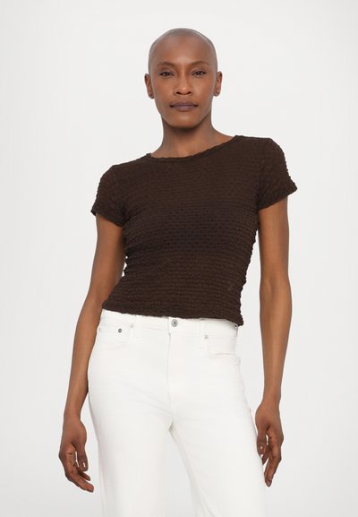 Tricou scurt cu mânecă, texturat, de culoare maronie, cu un decolteu rotund, asortat cu blugi albi. Tricoul are un aspect plisat sau zbârcit.