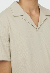 Camicia a maniche corte color beige chiaro con un motivo testurizzato, colletto a punta e vestibilità rilassata. I dettagli includono cuciture visibili sul colletto.