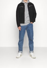 Chaqueta de ante negra con bolsillos de cremallera plateados, sudadera gris debajo, pantalones vaqueros azul enrollados y zapatillas blancas con detalles verdes.