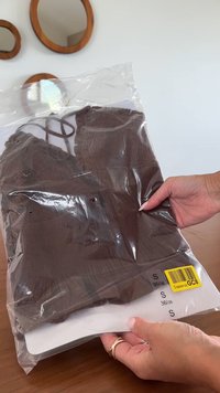 Hände halten ein kleines braunes, strukturiertes Oberteil in einer durchsichtigen Kunststoffverpackung mit Größenetiketten und einem gelben Barcode-Aufkleber.