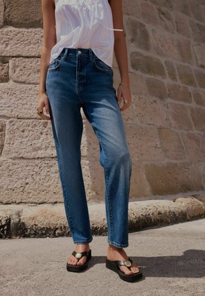 Person iført blå jeans med høj talje, hvid ærmeløs top, sorte platformsandaler, stående mod en stenmuren udendørs.