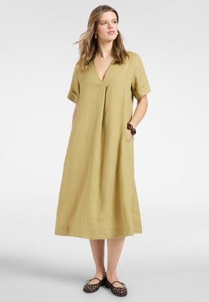 Frau trägt ein lockeres, knielanges senffarbenes Kleid mit kurzen Ärmeln, Taschen und schwarzen gewebten flachen Schuhen, steht mit einer Hand in der Tasche.