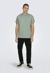 Man wearing rolled-sleeve sage green t-shirt, black pants, and black sneakers with white soles standing against plain light background.