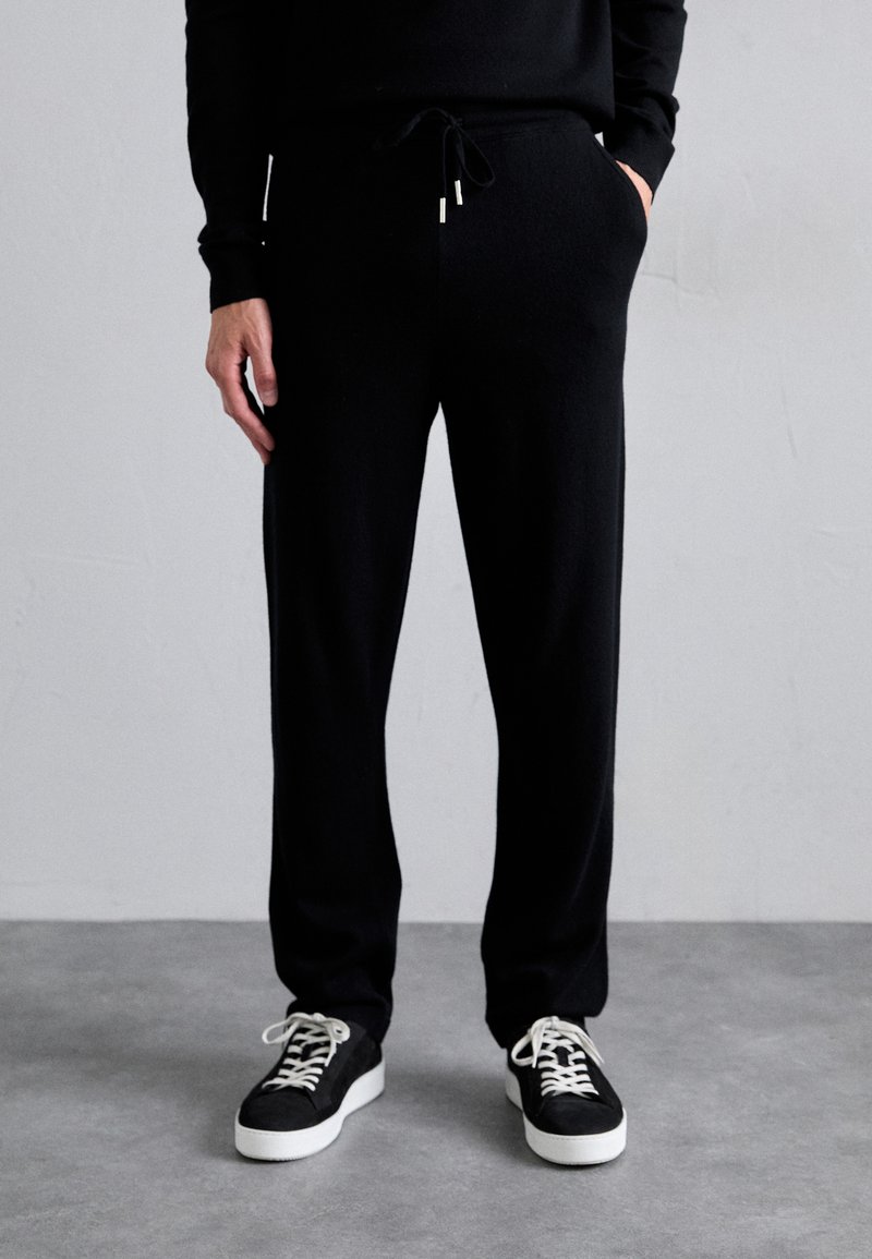 Black sweatpants with an elastic waistband and drawstring, featuring side pockets, paired with black and white sneakers on a gray background.