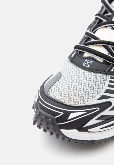 Κοντινό πλάνο της μύτης ενός ασπρόμαυρου αθλητικού παπουτσιού με πλέγμα ύφασμα, υφασμένη λαστιχένια σόλα και μαύρα κορδόνια σε λευκό φόντο.