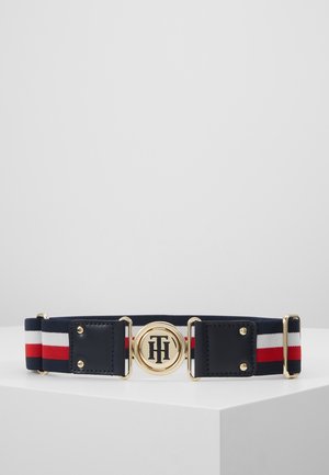 Pasek z tkaniny w paski w kolorach granatowym, białym i czerwonym z złotymi elementami i okrągłą złotą klamrą z wygrawerowanymi inicjałami na białej powierzchni.