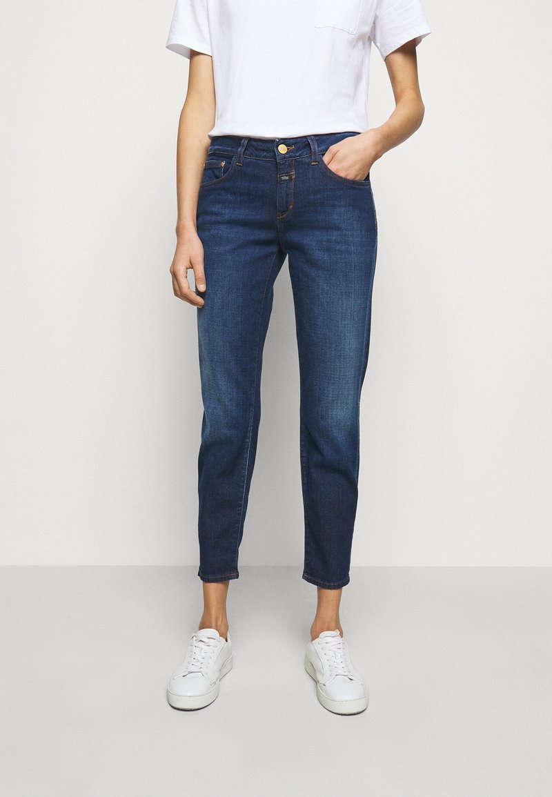 Femme portant un jean slim bleu foncé taille mi-haute, avec des détails de poche, des baskets blanches et un haut blanc à manches courtes.