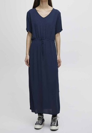 Kobieta ubrana w granatową midi sukienkę z dekoltem w serek, krótkimi rękawami i paskiem w talii, w połączeniu z czarno-białymi trampkami za kostkę.