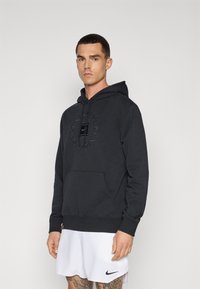 Black hoodie with a front pocket, featuring a tonal logo print. Material appears soft, with ribbed cuffs and hem, worn with white shorts.