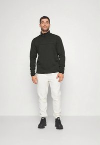 Melns vilnas pusmēteklis ar rāvējslēdzēju un teksturētu apdari, kombinēts ar baltām sporta biksēm un melniem sporta apaviem ar logo.