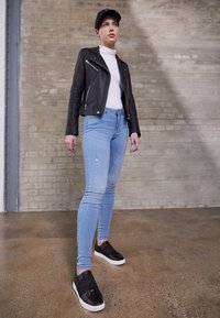 Veste de motard en cuir noir, col roulé blanc, jean skinny bleu clair avec détails usés, baskets noires à semelles blanches, et casquette noire.