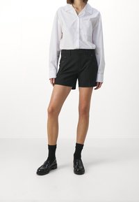 Shorts noirs ajustés avec des boutons croisés, associés à une chemise blanche à boutons. Le modèle porte des chaussettes noires et des mocassins brillants.