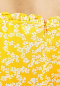 Kangas keltainen kukallinen, jossa on valkoisia kukkakuoseja, röyhelökaula-aukko ja näkyvä kultainen vetoketju takaosassa.