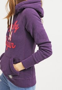 Sudadera con capucha morada y cordones blancos, texto bordado en rojo, bolsillo frontal y puños y dobladillo acanalados. Tejido de mezcla de algodón.