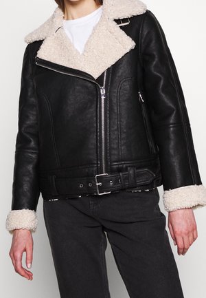 Schwarze Bikerjacke aus Kunstleder mit cremefarbenem Schaffellkragen und -manschetten, silbernem Reißverschluss und Gürtel, getragen zu schwarzen Hosen und weißem T-Shirt.