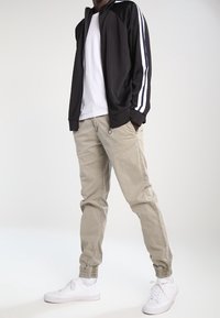 Svart zip-up jacka med vita ränder, vit t-shirt, beige byxor med elastiska ärmmuddar och vita sneakers, som visar en avslappnad, atletisk stil.