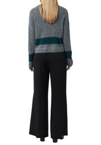 Kobieta z prostymi blond włosami ma na sobie szary sweter w tealowe paski oraz szerokie czarne spodnie, stoi plecami na białym tle.