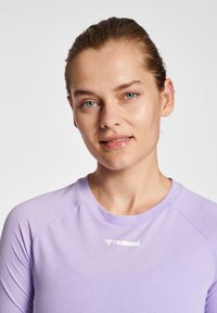 Jonge vrouw met blauwe ogen en gebonden haar, dragend een lichtpaars hummel sportshirt, kijkt licht glimlachend naar de camera.