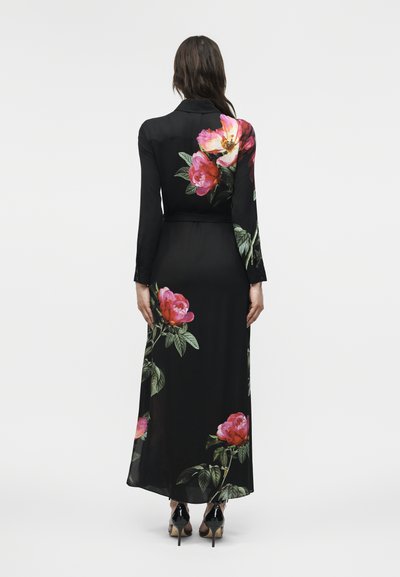 Mujer con un vestido largo de manga larga negro con grandes estampados florales en rosa y amarillo, de espaldas, llevando tacones negros.