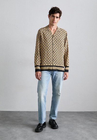 Beige lange mouwen shirt met een zwart geometrisch patroon, zonder kraag en met contrasterende zwarte manchetten, gecombineerd met lichtblauwe jeans.