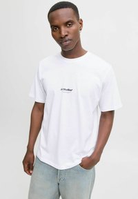 Camiseta de algodón blanca con un pequeño logo negro "JJ Studios*" en el pecho, mangas cortas y un corte relajado. Combinada con jeans de mezclilla azul claros.