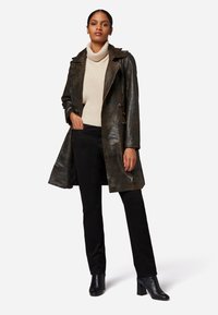 Brun läder trenchcoat med en vikbar krage, parat med en beige polokrage och svarta byxor. Modell bär svarta klackstövlar.