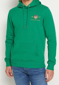 Vihreä huppari, valmistettu pehmeästä kankaasta, jossa on etutasku, säädettävä kiristysnyörillä varustettu huppu ja kirjailtu logo punaisena, sinisenä ja kultaisena.