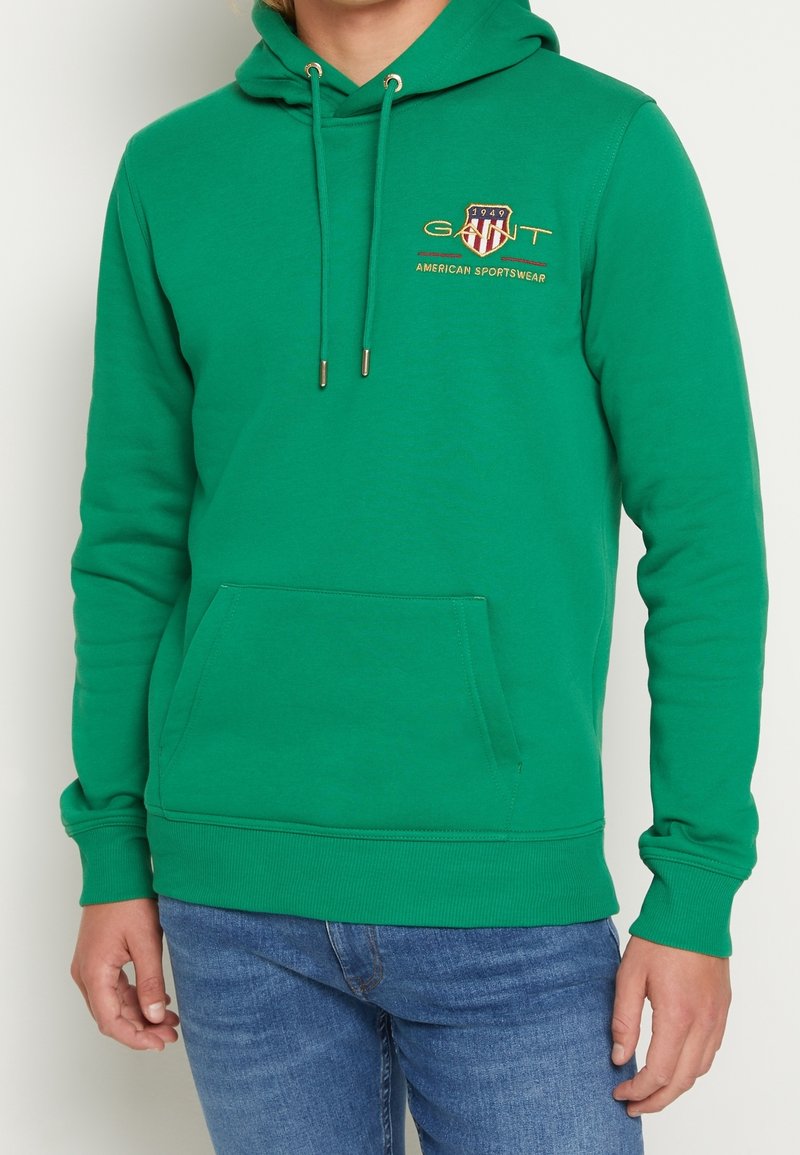 Vihreä huppari, valmistettu pehmeästä kankaasta, jossa on etutasku, säädettävä kiristysnyörillä varustettu huppu ja kirjailtu logo punaisena, sinisenä ja kultaisena.