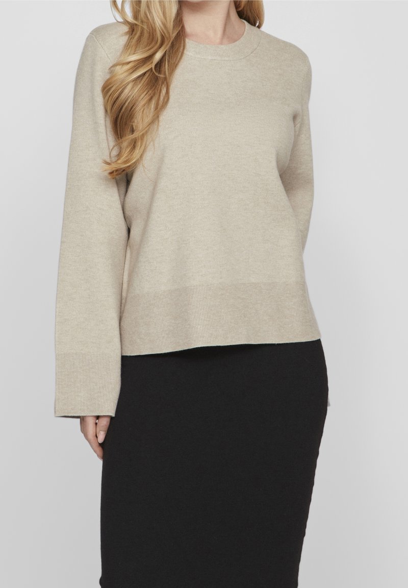 Beige Pullover mit Rundhalsausschnitt, langen Ärmeln und gerippten Abschlüssen, mit lockerer Passform und weicher Textur. Getragen über einem schwarzen Rock.