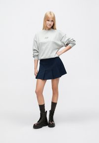 Γκρι cropped φούτερ με λογότυπο, σκούρο μπλε πλισέ φούστα, καφέ κάλτσες μέχρι το γόνατο και μαύρα αρβυλάκια με κορδόνια και χοντρές σόλες.