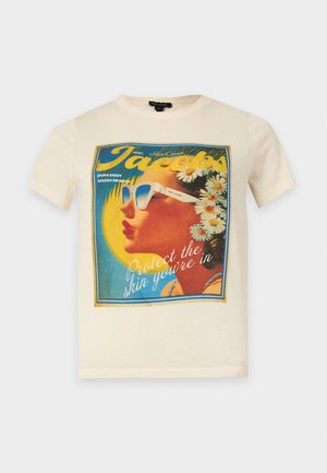 Femme debout, portant un t-shirt beige à motif avec texte, jean bleu évasé, chaussures pointues, main dans la poche, sur fond uni.