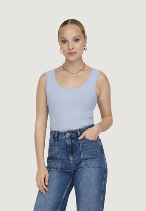 Donna con capelli biondi raccolti, che indossa orecchini a cerchio dorati, una collana a catena, un top azzurro senza maniche e jeans blu a vita alta, con la mano in tasca.