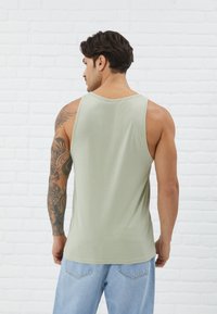 Light green tank top made of smooth fabric, featuring a classic cut with wide armholes. Worn with light blue denim jeans.