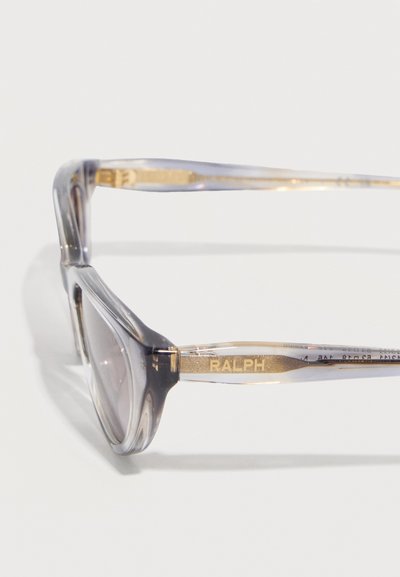 Διαφανή γκρι πλαίσια γυαλιών με καθαρούς φακούς και χρυσές λεπτομέρειες στους βραχίονες, με έναν βραχίονα που φέρει τη λέξη "RALPH" με χρυσά γράμματα.