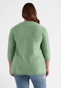 Groene gebreide trui met driekwartmouwen, voorzien van een gestructureerd patroon en subtiele sprankels, gecombineerd met een blauwe jeans.