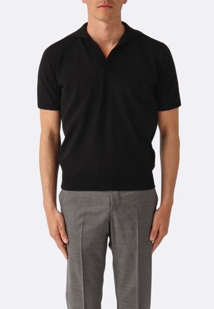 Uomo che indossa una polo a maniche corte nera in maglia con pantaloni eleganti grigi a pieghe, in piedi contro uno sfondo chiaro e uniforme.