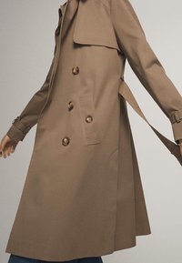 Trench-coat beige croisé à quatre boutons, poches latérales et ceinture, porté par une personne avec les bras légèrement étendus.