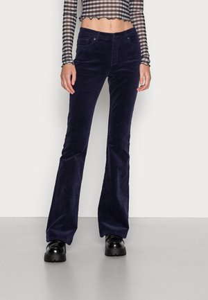 Kvinde iført højtaljede mørkeblå manchesterflared bukser, sorte chunky platformsko og en langærmet sort-hvid ternet cropped top.