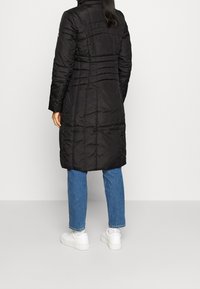 Svart lång pufferjacka med hög krage och stickad detaljer på ryggen, kombinerad med blå jeans och vita sneakers.