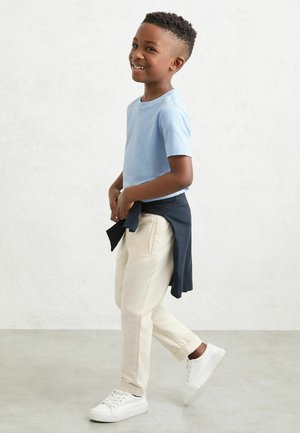 Băiat zâmbind și mergând, purtând un tricou albastru deschis, pantaloni bej, adidași albi și o jachetă închisă la culoare legată în jurul taliei.