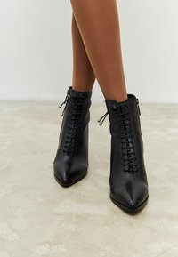 Botas negras de cuero hasta el tobillo con punta afilada, cordones frontales y detalle de cremallera lateral. Textura suave, tacón plano, adecuadas para diversos atuendos.
