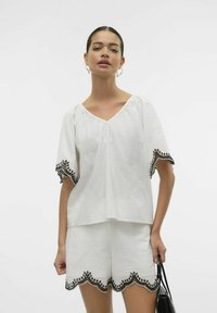 Top bianco in lino con maniche corte, caratterizzato da accenti ricamati neri su maniche e orlo, abbinato a pantaloni corti coordinati.