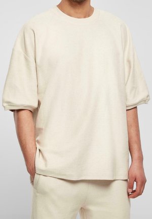 Man wearing loose, cream-colored short-sleeve textured top and matching pants, standing with one hand in pocket.