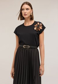 Sort kortærmet top med et blomstret udskæringsdesign og sorte detaljer. Sammen med en plisseret sort nederdel og et bælte med guldspænde.
